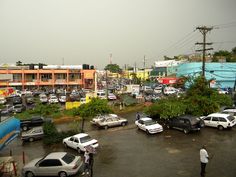 a parking lot filled with lots of cars next to tall buildings and people walking on the sidewalk