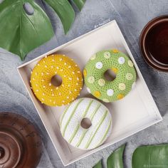 three decorated donuts in a box next to some green leafy leaves and a cup of coffee