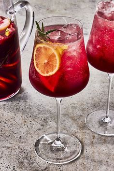 two wine glasses filled with red liquid and garnished with orange slices next to a pitcher