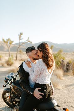 a man and woman sitting on a motorcycle in the desert, kissing each other's foreheads