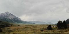 an open field with mountains in the background and trees on the other side, under a cloudy sky