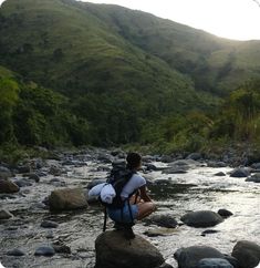 a person sitting on rocks in the middle of a river with mountains in the background