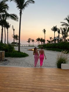 two women in pink dresses walking on the beach at sunset with palm trees behind them