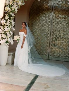 a woman in a wedding dress standing next to a flower covered archway with white flowers