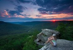 a woman sitting on top of a large rock next to a lush green forest under a cloudy sky