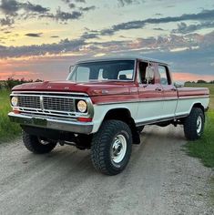 an old red pickup truck parked on the side of a dirt road at sunset with clouds in the background