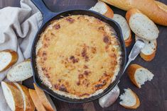 a skillet filled with cheese and bread on top of a wooden table next to rolls