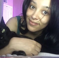 a woman is holding a black cat in her arms and smiling at the camera while sitting on a bed