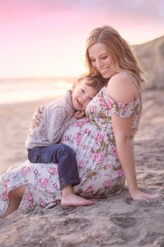 a woman holding a child on top of a sandy beach next to the ocean at sunset