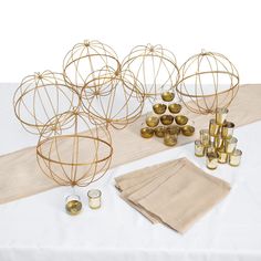 a table topped with lots of gold vases next to napkins and candles on top of a white cloth covered table