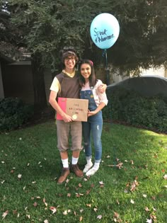 a man and woman standing in the grass holding a box with a balloon attached to it