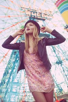 a woman standing in front of a ferris wheel with her hands on her head and wearing a hat