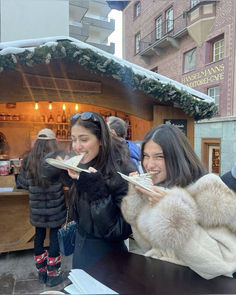 two women are eating food outside in the snow while people look on from behind them