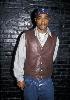 a man standing in front of a brick wall wearing a leather vest and bandana