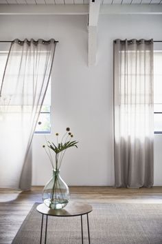 a vase with flowers sitting on top of a table in front of two window curtains