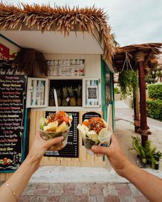 two people holding up food in front of a small building with tiki hut on the side