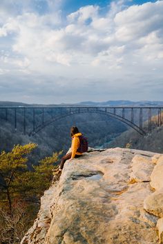 a person sitting on top of a large rock next to a bridge and looking at the sky