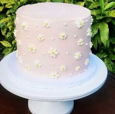 a pink cake with white flowers on it sitting on a table next to some bushes