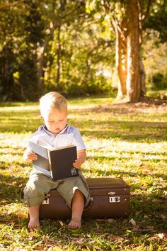 a young boy sitting on top of a suitcase holding a book in his hands and looking at it