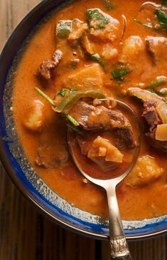 a blue bowl filled with stew and meat on top of a wooden table next to a spoon