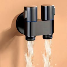 two black faucets with water coming out of them in front of a tan wall