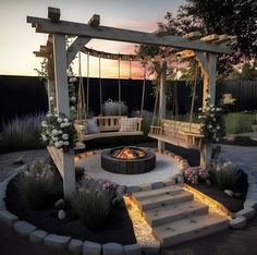 an outdoor fire pit with swings and seating around it at dusk or dawn, surrounded by flowers and greenery