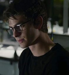 a young man wearing glasses looking at his cell phone in the dark room with other items on the table behind him