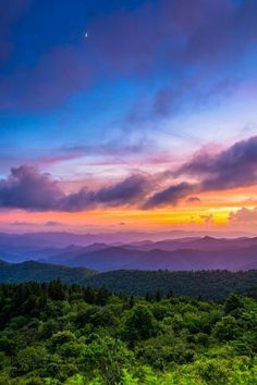 the sun is setting over mountains and trees in the foreground, with clouds above