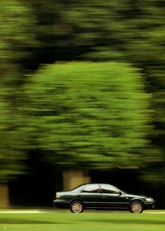 a black car driving down a street next to tall green grass and trees in the background