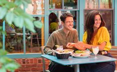 a man and woman sitting at a table with food in front of them smiling for the camera