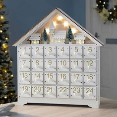 a white wooden house shaped calendar with christmas lights on the front and sides, sitting on a table next to a tree