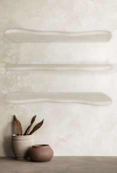 two white vases sitting on top of a table next to a plant in front of a wall