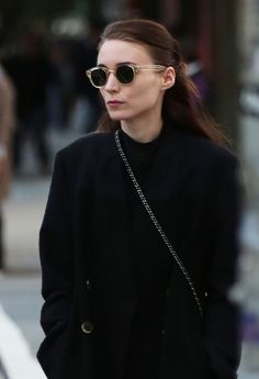 a woman in black coat and sunglasses walking down the street with her hand on her hip