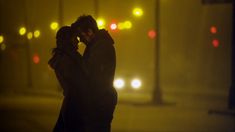 a man and woman kissing in the street at night with traffic lights behind them on a foggy day