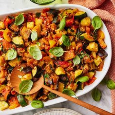 a white dish filled with cooked vegetables and topped with basil leaves next to a wooden spoon