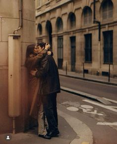 two people standing next to each other on the side of a street with buildings in the background