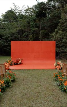 an outdoor ceremony setup with chairs and flowers on the grass, in front of a red backdrop