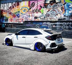 a white car with blue rims parked in front of a graffiti covered wall,