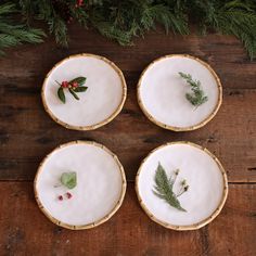 four plates with plants on them sitting on a wooden table next to evergreen leaves and berries