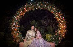 a man and woman sitting on top of a bench in front of a floral arch