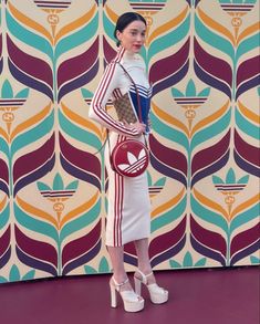 a woman standing in front of a colorful wall holding a purse and wearing white shoes