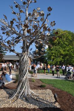 a metal tree sitting on top of a pile of dirt next to a park filled with people