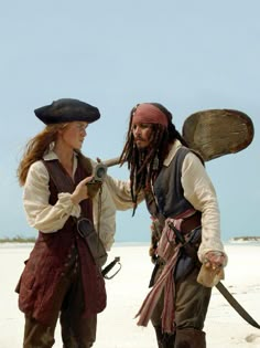 two people dressed in pirate costumes standing on the beach, one holding a camera and looking at another person's face