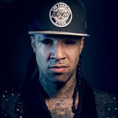 a tattooed man wearing a black hat and jacket with tattoos on his arm, looking at the camera