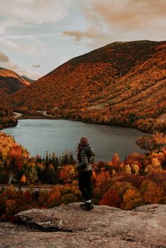 a person standing on top of a mountain overlooking a lake and trees with fall colors