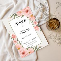 a wedding card with pink flowers and greenery next to it on a white blanket