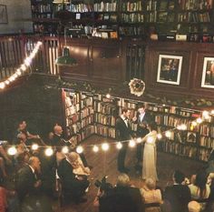 a couple is getting married in front of a large group of bookshelves with string lights strung across the room