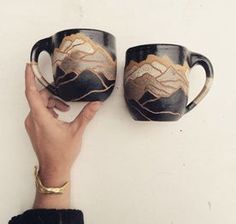 two coffee mugs with mountains painted on them are held up against a white wall