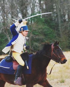 a man riding on the back of a brown horse in a field with trees behind him