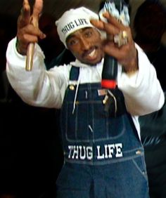 a man in overalls and a hat holding up two hotdogs to the camera